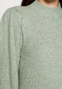 Jasnozielony, prążkowany sweter z wysokim dekoltem i bufkami na ramionach, ukazujący miękką teksturę i pionowy wzór.