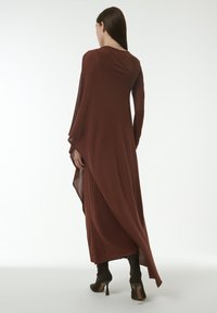 Robe asymétrique marron à manches longues, confectionnée dans un tissu lisse. Présente des détails transparents sur les manches et des bottes noires à talons hauts.