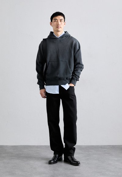 Sudadera con capucha gris oscuro y bolsillo frontal, llevada sobre una camiseta blanca, combinada con pantalones negros y zapatos negros. Diseño simple y casual.
