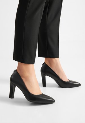 Mujer con zapatos de tacón altos negros de punta puntiaguda combinados con pantalones negros recortados, de pie sobre una superficie blanca.