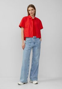 Czerwona koszula z krótkim rękawem i kołnierzykiem, w parze z niebieskimi spodniami typu wide-leg. Strój charakteryzuje się luźnym krojem, gładkim materiałem i prostymi detalami projektowymi.