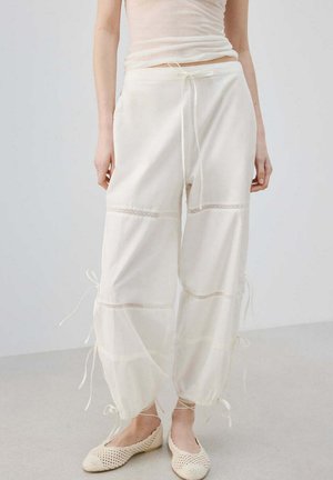 Kobieta nosząca luźne, przezroczyste białe spodnie ze sznurkami w pasie, po bokach i przy kostkach, w połączeniu z plecionymi kremowymi balerinami.