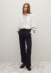 Weiße Bluse mit langen Ärmeln und braunen Steppnähten, kombiniert mit dunkelblauen, ausgestellten Jeanshosen und schwarzen klobigen Schuhen, in einem neutralen Ambiente.