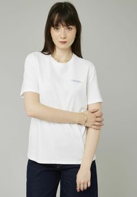 T-shirt en coton blanc avec un col rond et des manches courtes, comportant un texte bleu clair et jaune sur le côté gauche de la poitrine.