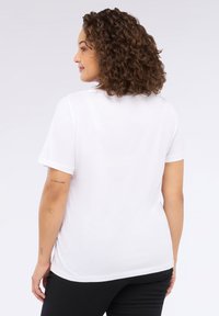 Femeie cu păr creț, îmbrăcată cu un tricou alb simplu cu mânecă scurtă și pantaloni negri, stând cu spatele, arătând tatuaje pe brațul ei stâng.