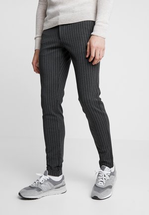 Person iført mørkegrå, smalle bukser med striber, lys beige sweater og grå og hvide sneakers, stående på en ensfarvet baggrund.