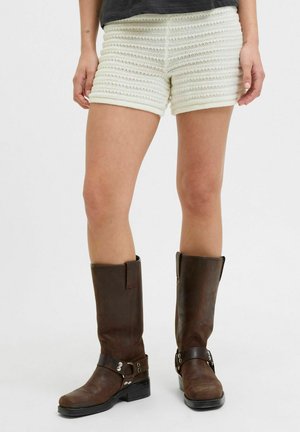 Shorts de punto blanco texturizado con rayas horizontales, combinados con botas de cuero marrón hasta la rodilla que presentan un detalle de hebilla y suelas planas.