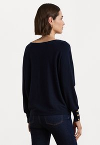 Tengerés kék kötött pulóver széles nyakkivágással, ejtett vállakkal és bordázott mandzsettákkal, három aranyszínű gombdíszítéssel.