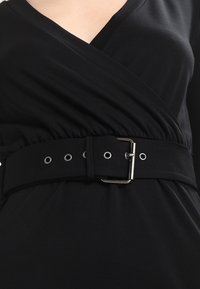 Černé šaty s překříženým designem a nastavitelným páskem se stříbrnou sponou a očky, vyrobené z měkké, elastické tkaniny.