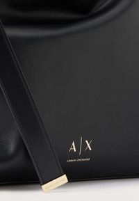 Μαύρη τσάντα Armani Exchange με χρυσό λογότυπο A|X και μαύρο λουράκι με χρυσές λεπτομέρειες, πάνω σε λευκή επιφάνεια.