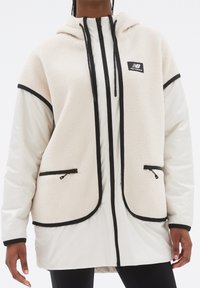 Krämfärgad fleecejacka och vit nylonjacka med huva, med svarta detaljer och fickor med dragkedjor. Texturerad kropp och släta ärmar.