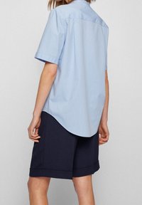Himmelblaues Kurzarmhemd mit abgerundetem Saum und Rückenpasse, kombiniert mit dunkelblauen Shorts mit umgeschlagenem Bund.