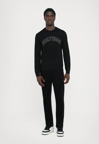 Sort langærmet skjorte med "VOLTAIRE" trykt i buet hvid tekst, parret med sorte bukser og sort-hvide sneakers. Enkel design.