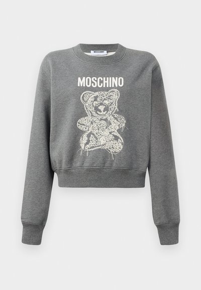 Pelēks īsais sporta krekls ar baltu grafisku lāča dizainu, virs kura atrodas teksts "MOSCHINO". Mīksta tekstūra un ribotas aproces pie piedurknēm.