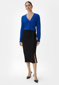 Blauwe gebreide cardigan met een V-hals en een gestructureerd patroon, gecombineerd met een zwarte midi-rok met een zijsplit en strikdetail. Zwarte hoge hakken.