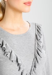 Pull en gris avec un col rond et des accents de volants verticaux sur le côté gauche. Texture tricotée douce et design ajusté.