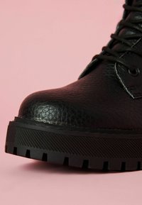 Černá kožená bota s texturovaným povrchem, kulatou špičkou a silnou černou gumovou podrážkou s vroubkovaným vzorem. Pozadí v jemné růžové.