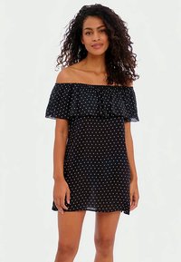 Mulher com cabelo encaracolado vestindo um vestido preto de ombros à mostra com bolinhas brancas, de pé contra um fundo branco liso.