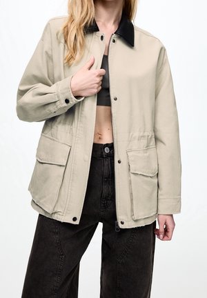 Frau trägt eine beige Jacke mit schwarzem Kragen über einem schwarzen kurzen Oberteil und hoch taillierten schwarzen Jeans vor weißem Hintergrund.