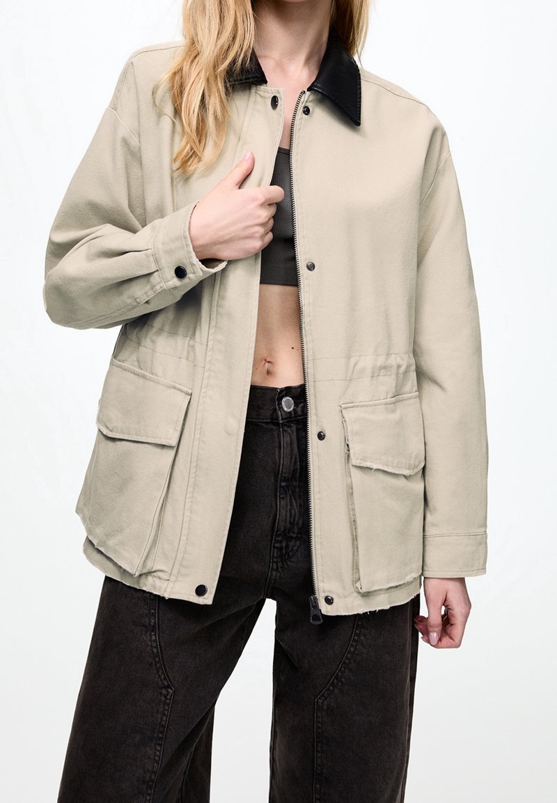 Femme portant une veste beige avec un col noir sur un top court noir et un jean noir taille haute sur fond blanc.
