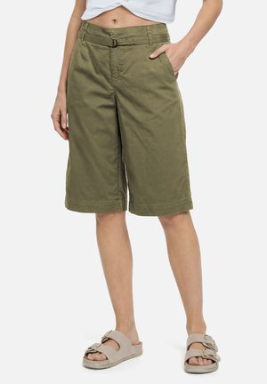 Frau trägt olivgrüne knielange Shorts mit Gürtel und beige Slip-on-Sandalen, Hand in der Tasche, vor weißem Hintergrund.