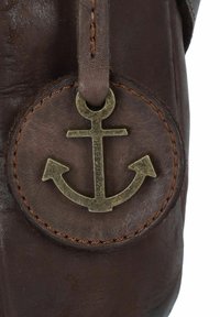 Detalle de un bolso de cuero marrón que presenta una etiqueta metálica redonda con un emblema de ancla y costuras beiges a lo largo del borde exterior.