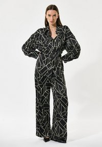 Črn in bež vzorčast romper s kratkimi rokavi in širokimi hlačami, ki ima geometrijski vzorec in gladko satensko teksturo.