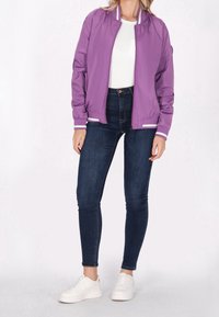 Femme portant une veste bomber violette, un t-shirt blanc, des jeans skinny foncés et des baskets blanches, debout devant un fond uni.