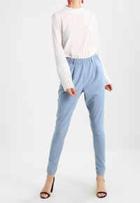 Un haut blanc à manches longues avec des poignets plissés, associé à un pantalon tapered bleu clair. Des sandales à talons noirs complètent le look.