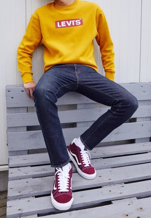 Osoba ubrana w musztardowożółtą bluzę Levi’s, ciemnoniebieskie dżinsy oraz czerwono-białe sneakersy, siedząca na jasnoszarej, drewnianej ławce.