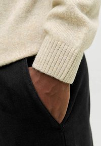 Beige gebreide trui met geribbelde manchetten, gecombineerd met zwarte broek. Close-up met hand in zak, waarbij textuur en materiaalcontrasten worden getoond.