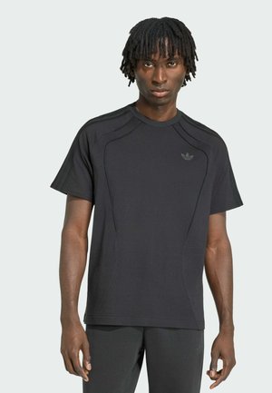 Junger Mann mit kurzen Dreadlocks, der ein schwarzes Adidas-T-Shirt und dunkle Hosen trägt, steht vor einem einfarbigen hellgrauen Hintergrund.