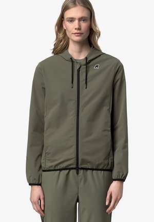 Donna che indossa una giacca verde oliva con zip e cappuccio e pantaloni abbinati, rivolta frontalmente con espressione neutra su uno sfondo chiaro e uniforme.