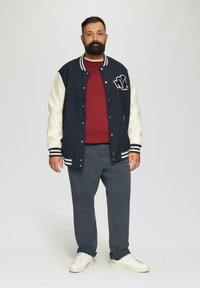 Marineblauwe en crèmekleurige varsityjack met gestreepte boorden, rode trui, grijze broek en witte sneakers. Kenmerkt zich door logo op het jack. Casual pasvorm.