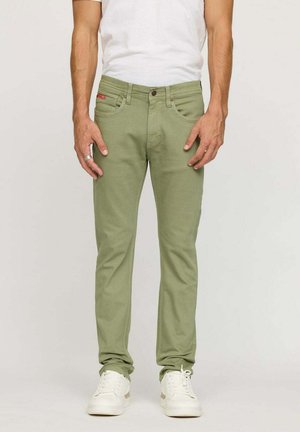 Jean en denim vert olive avec une coupe slim, cinq poches et une étiquette logo rouge. Associé à des baskets blanches sur un fond uni.
