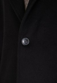 Cappotto di lana nero con rever a punta e chiusura a un bottone. Il bottone è nero con quattro fori. Il tessuto ha una consistenza morbida.