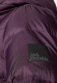 Paarse pufferjack-fabric met een textuurfinish. Een zwart vierkant label met het reliëf van het 'Jack Wolfskin'-logo is zichtbaar op de mouw.