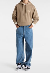 Sweat à capuche marron avec poche frontale et logo ; jean ample bleu clair ; baskets noires et blanches. Coupe décontractée avec texture douce.