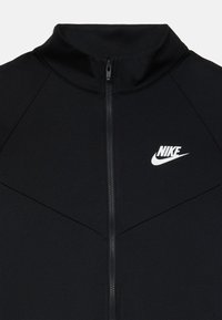 Černá zipová bunda vyrobená z hladké tkaniny, s vysokým límcem a bílým logem Nike na hrudi, s minimalistickými designovými liniemi.