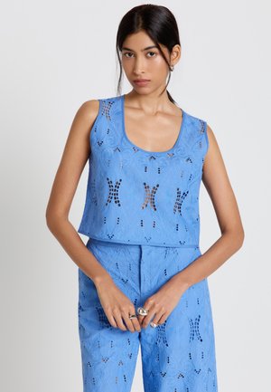 Femme aux cheveux foncés portant un haut court en œillet bleu sans manches et un pantalon taille haute assorti, posant avec les mains jointes devant elle.