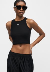 Γυναίκα που φοράει μαύρο αμάνικο crop top με το λογότυπο της Adidas, μαύρο αθλητικό παντελόνι και μεγάλα μαύρα γυαλιά ηλίου σε απλό φόντο.