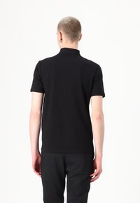 Juodas trumpas rankovės polo marškinėliai su aukštu apykakle, lygiu paviršiumi ir siauru siluetu. Modelis parodytas iš nugaros, vilkintis juodas kelnes.