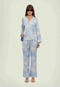 Naine kannab sinist pilvemustriga pidžaamastiilis ülikonda, musti päikeseprille, beeži sandaale ja tal on kaasas sinine õlakott.