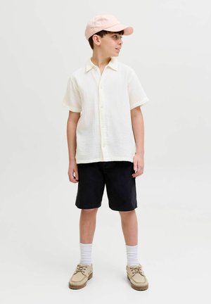 Zēns, kurš valkā gaiši rozā cepuri, teksturētu īsroku baltu kreklu ar pogām, melnas bikses šortus, baltas zeķes un bēšas kurpes, skatās uz sāniem.