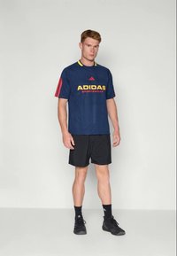 Mėlyna Adidas sportinė marškinė su geltonu logotipu, raudonomis juostomis ant rankovių, derinama su juodomis šortais ir juodais bateliais, vilkima stovinčio vyriško modelio.
