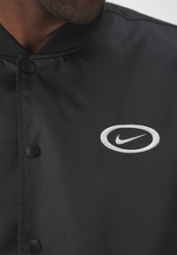 Černá bunda s hladkou, lesklou texturou, s bílým vyšitým logem Nike. Má zapínání na knoflíky a žebrovaný límec.