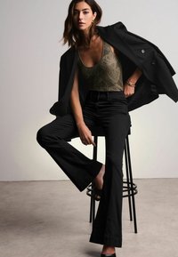 Jeans neri a palazzo abbinati a un top in pizzo verde oliva indossato sotto un blazer nero, che mette in risalto una texture liscia e un design sartoriale.
