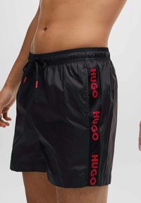 Čierne plavky z hladkého materiálu s elastickým pásom a šnúrkou. Má červené logo "HUGO" pozdĺž strany.
