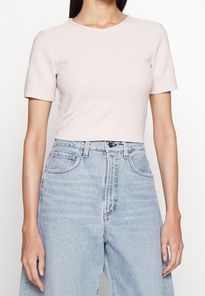 Vrouw draagt een geribd, lichtroze kort mouw crop top en hooggetailleerde lichtblauwe spijkerbroek, staat met ontspannen handen.