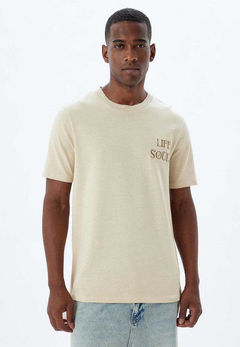 Beige kortemouwen t-shirt van katoen met een ronde hals, voorzien van bruine geborduurde tekst "LIFE" en "SOUL" op de linkerborst.